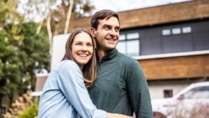 A married couple smiles in front of a modern home.