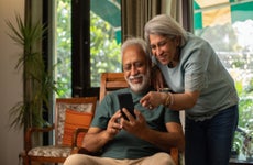 Smiling senior woman standing and pointing at smart phone being used by an older man sitting on chair at home.