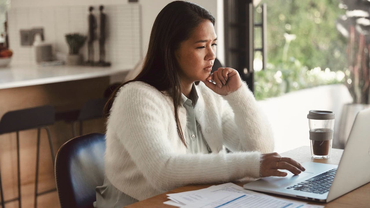 The woman sits at her dining table with her computer, carefully examining bills. The backdrop is her kitchen, and the atmosphere is tense as she contemplates her financial situation.