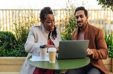 A multi-racial couple looks at a laptop while having coffee in a rooftop garden.