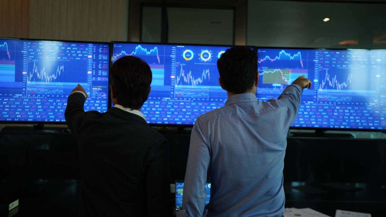 Two traders watch stocks markets on three screens