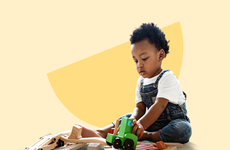 A toddler wearing overalls plays with toys in front of a yellow background.