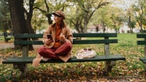 Beautiful mid adult woman sitting on bench and using mobile phone outside in the city on autumn day.