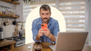 A white man sits at a kitchen table while using an orange phone. There is a laptop open in front of him. Design includes a light yellow semicircle behind him.