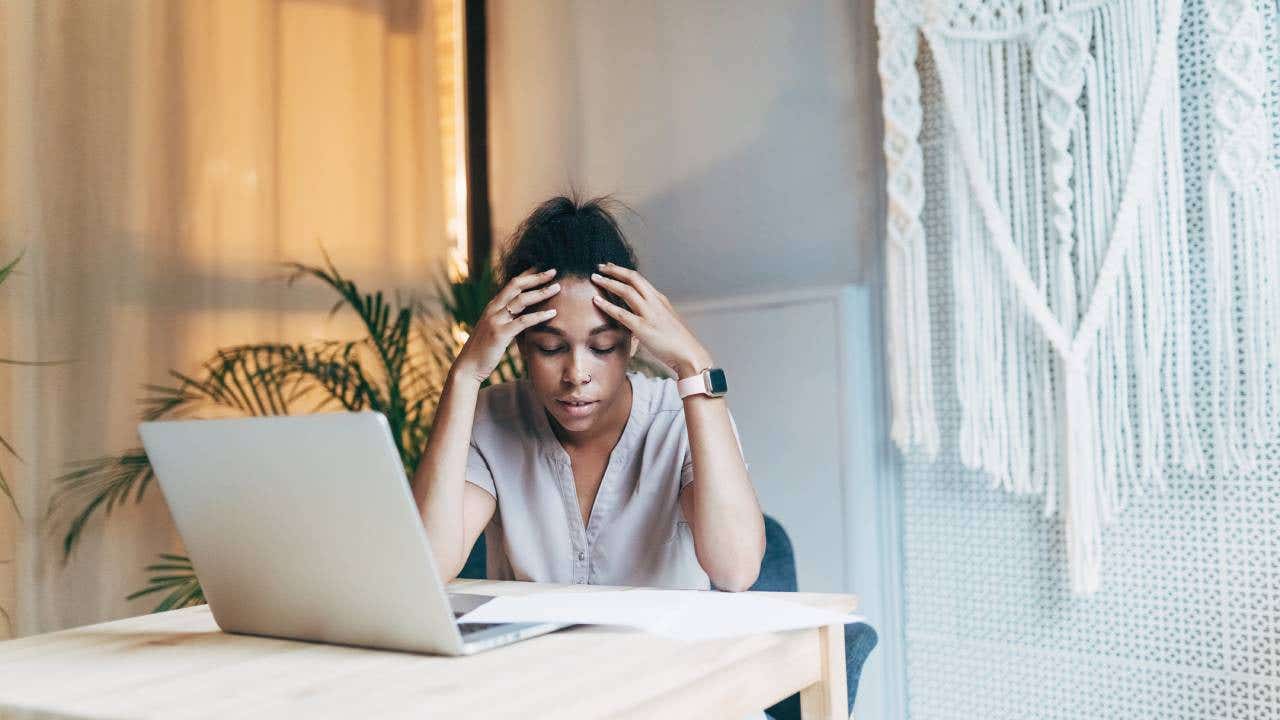 A young mixed race woman sits at her laptop computer looking stressed with her head in her hands.