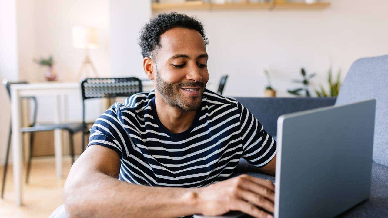 Young adult man using laptop computer while resting on sofa at home.