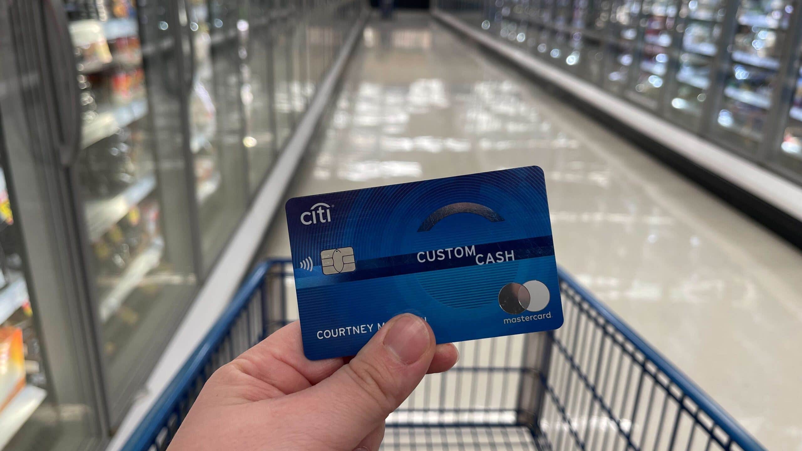 Closeup of a person holding a credit card over a grocery cart in a frozen food aisle.