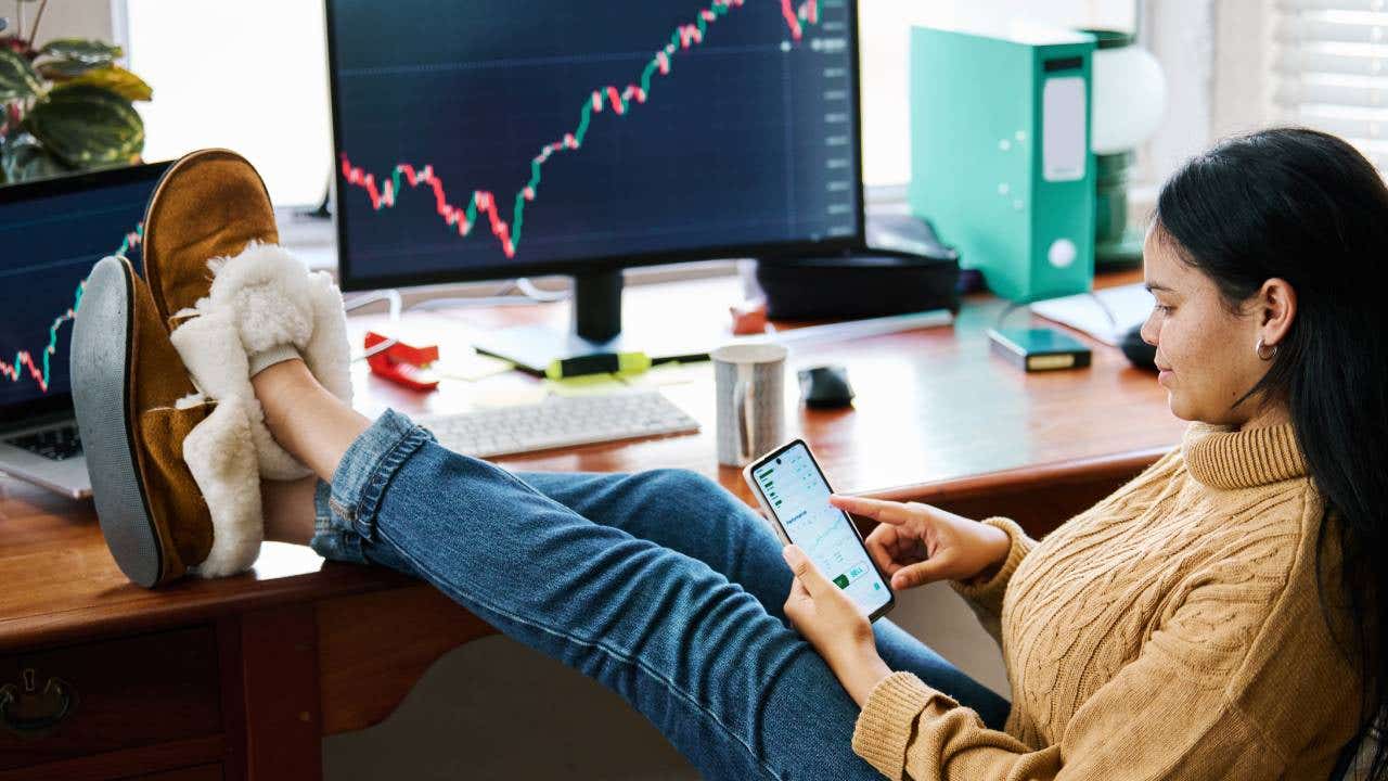 Young woman using a smartphone app to manage and analyze stock market data while sitting at a home office with her feet up on the desk