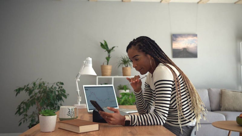 Person looking down at their smartphone while sitting next to a laptop displaying a stock chart.