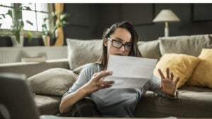 Woman in glasses seated in her living room reviewing a paper near her laptop