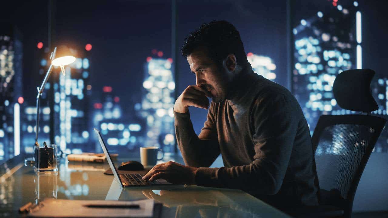 A man looks at his laptop at night with a lit up cityscape behind him.