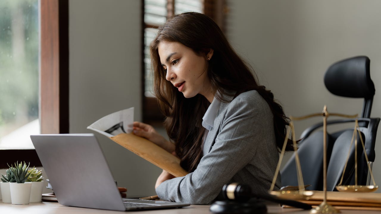 Young Asian woman in professional sits at a desk with a gavel and scales on it. She is speaking while looking through papers.