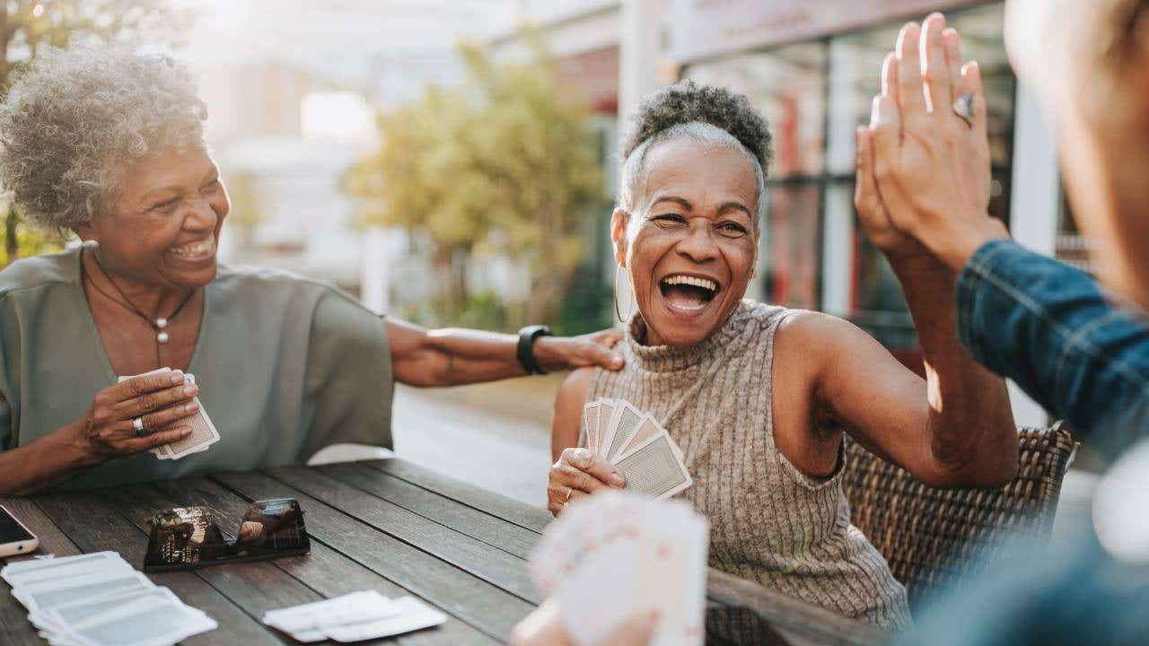 Senior women play cards outside, high-fiving and laughing together.