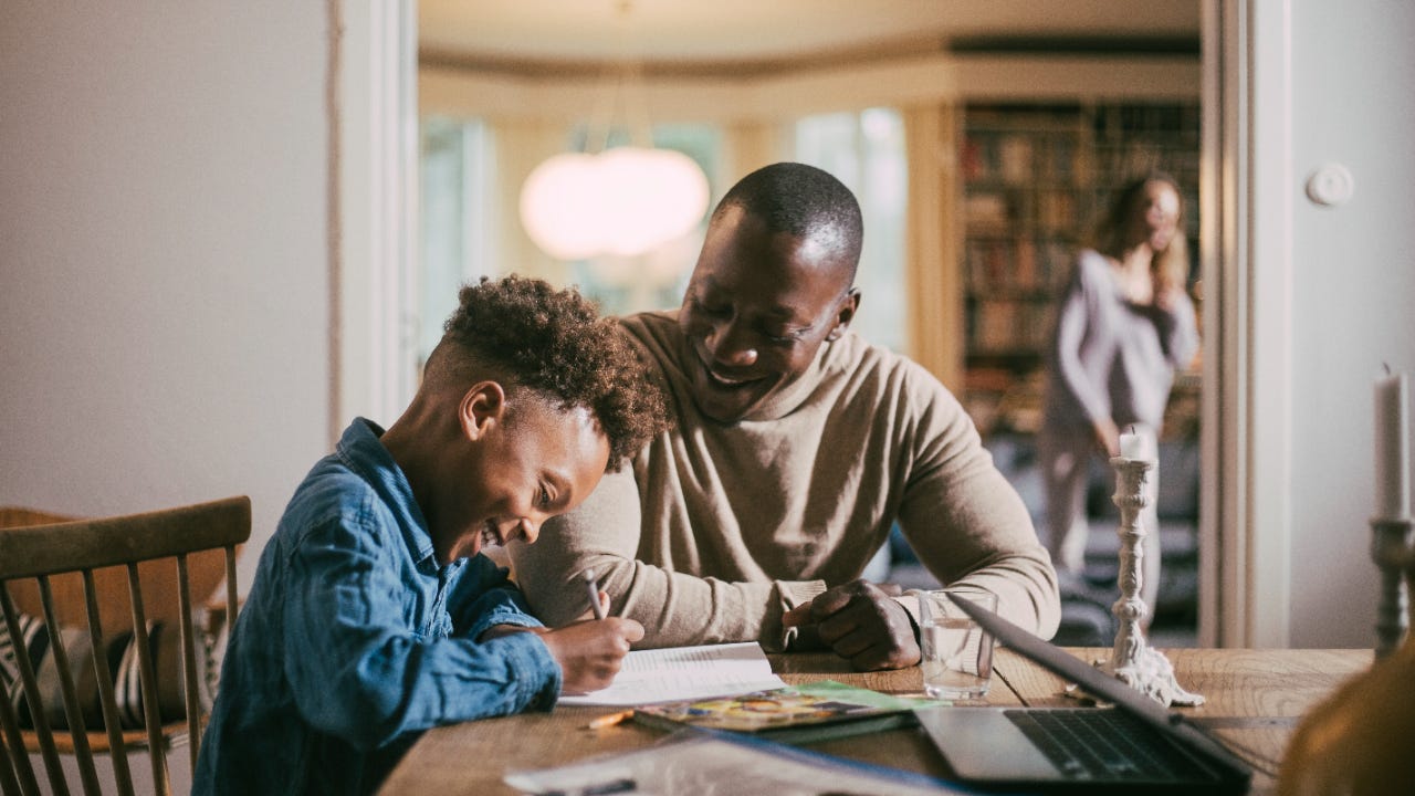 A Black man smiles brightly as he helps his young Black son with homework. They are sitting together at their dinner table.