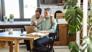 A mature man works on his laptop in the kitchen while his wife is next to him.