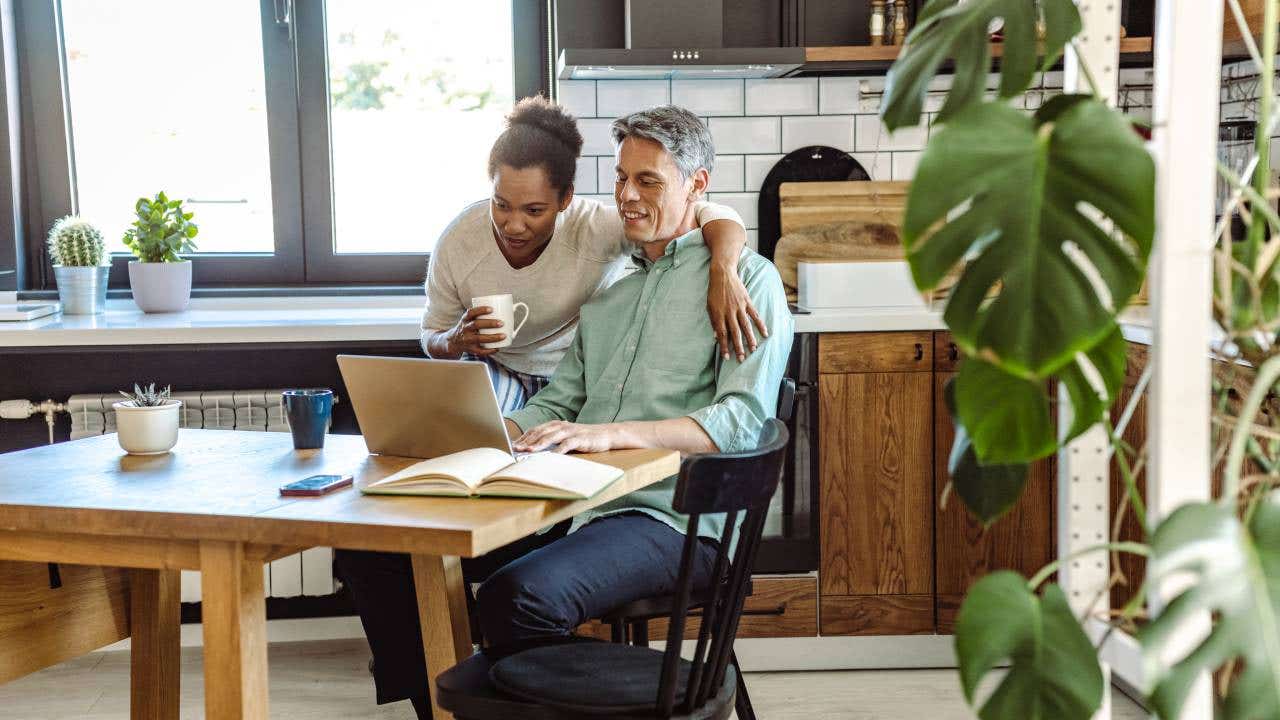 A mature man works on his laptop in the kitchen while his wife is next to him.