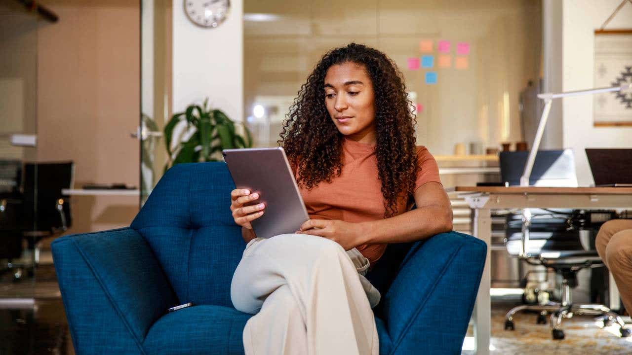 A young businesswoman looks at her tablet while sitting on an armchair at an office.