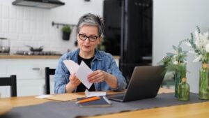 Serious mature woman at a table in a home interior looks through mail and bank accounts.