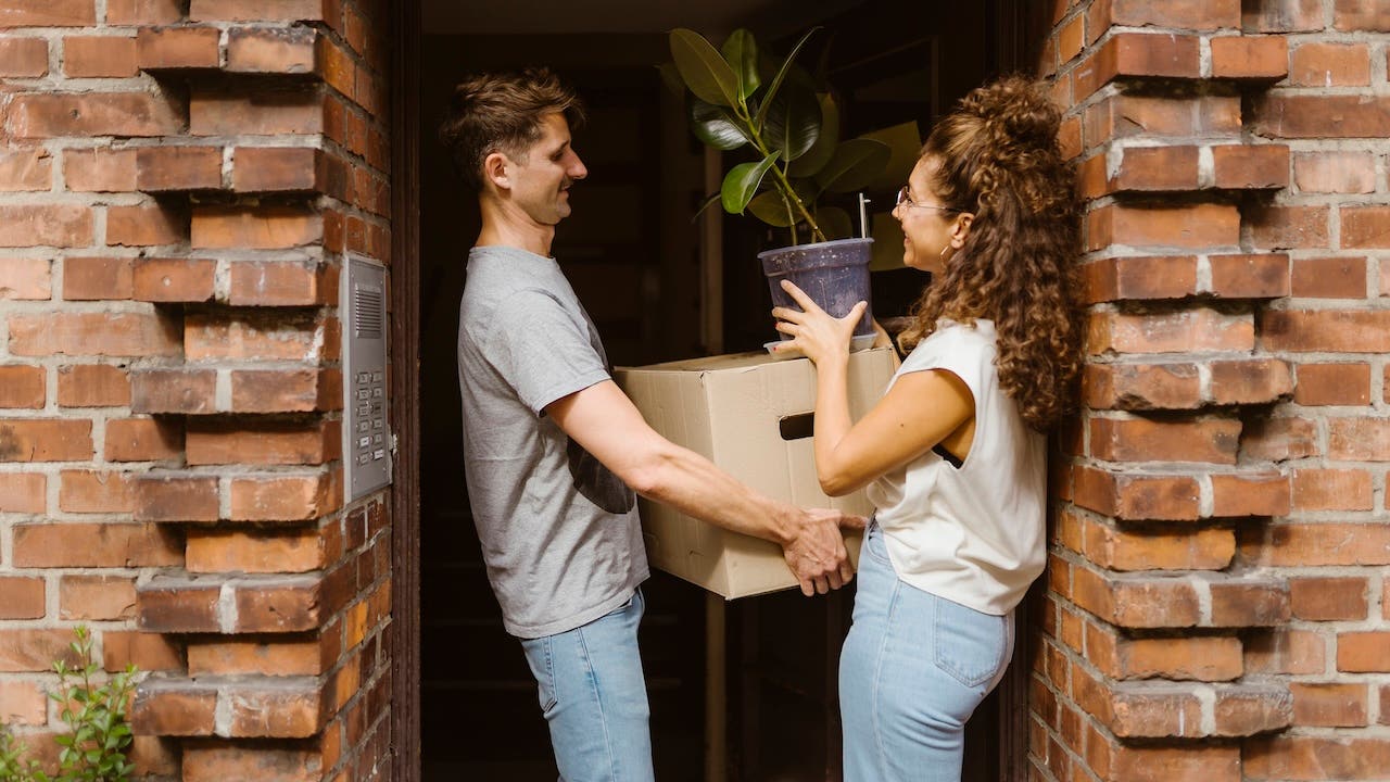 Happy couple with cardboard box and potted plant talking to each other at doorway