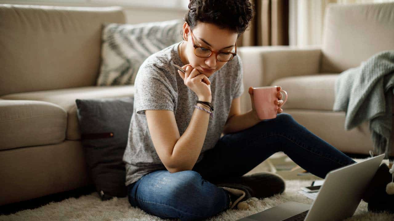 A young woman sits on the floor, holding a mug and a pen, looking at her laptop on the floor.