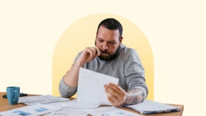 Man thoughtfully reviewing a document at a table that has many papers on it. The image has a light yellow background with a darker yellow circle behind the man.