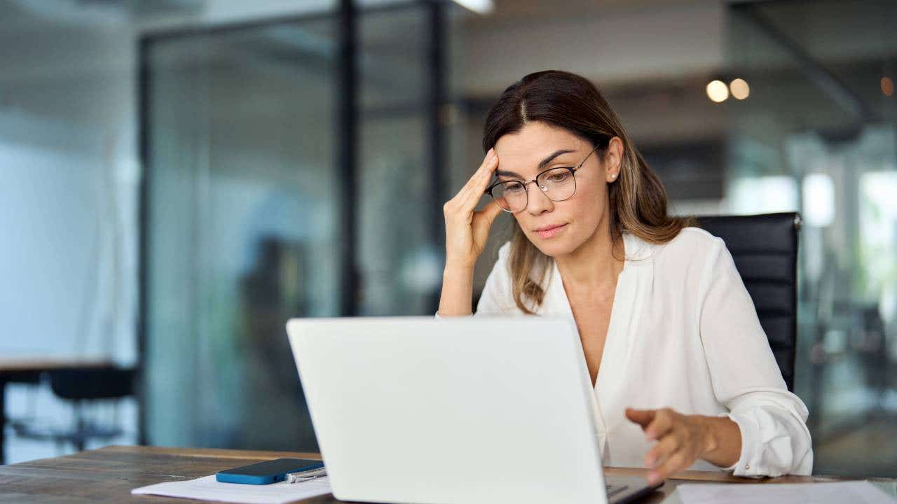 Woman with glasses in front of a computer with her hand by her head, looking worried.