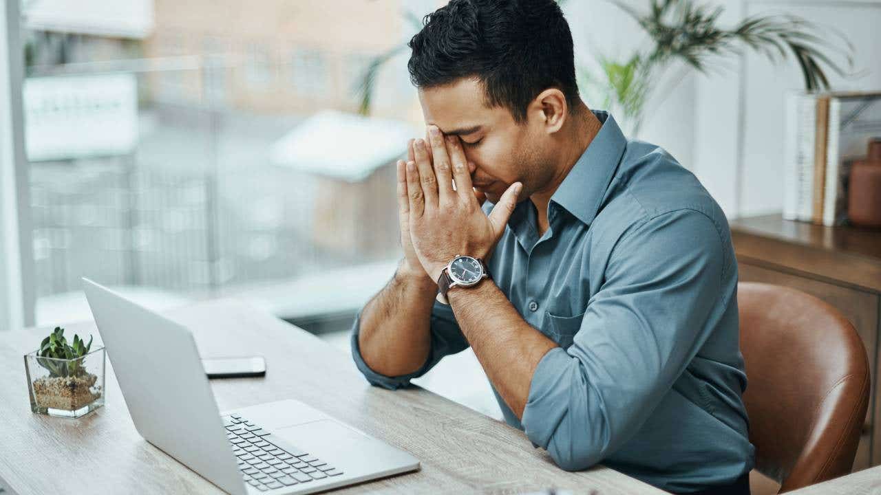 A man sits at a desk with a laptop computer and his hands by his face in an expression of stress.