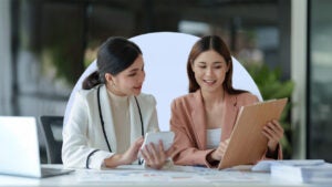 Two smiling Asian women dressed in business attire review documents on a clipboard while working in a cafe. The image includes an off-white half circle behind the two women.