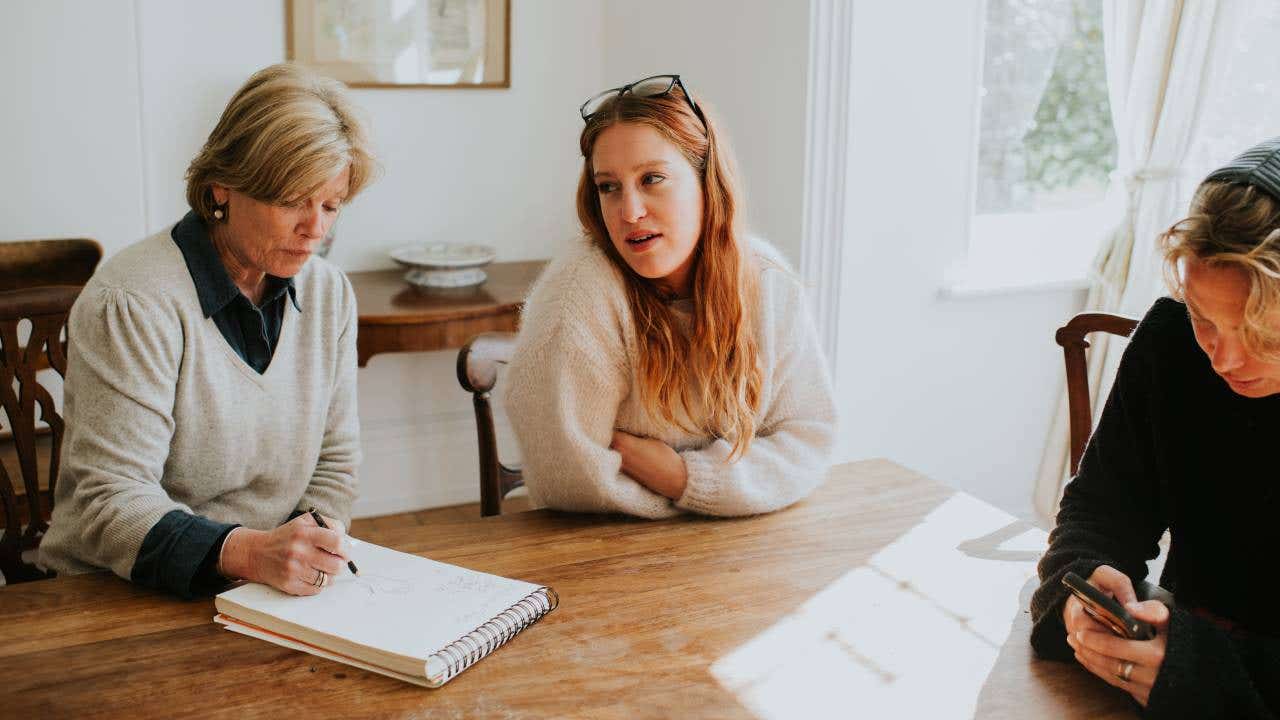 Two younger woman sit around a dining table with an older woman who takes notes on a notepad.