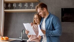 A smiling young couple looks at paperwork in a kitchen.