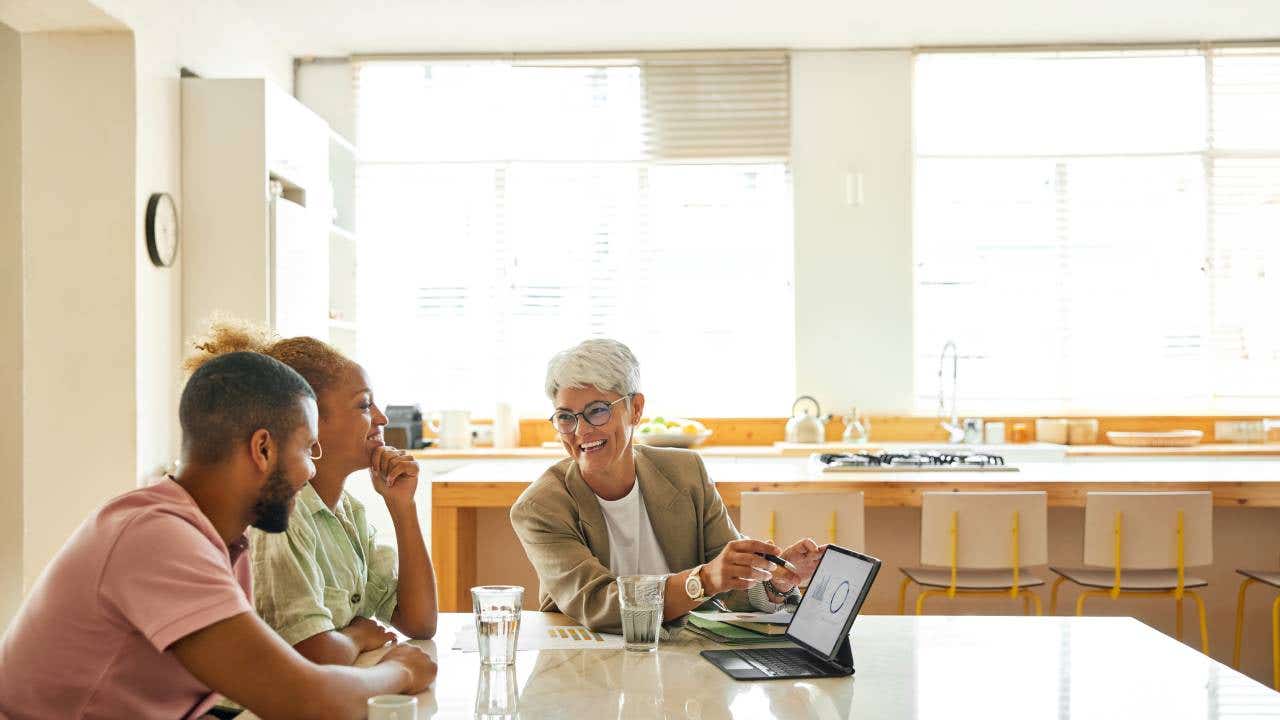 A couple sits at a table while a financial advisor points to a tablet displaying some charts.