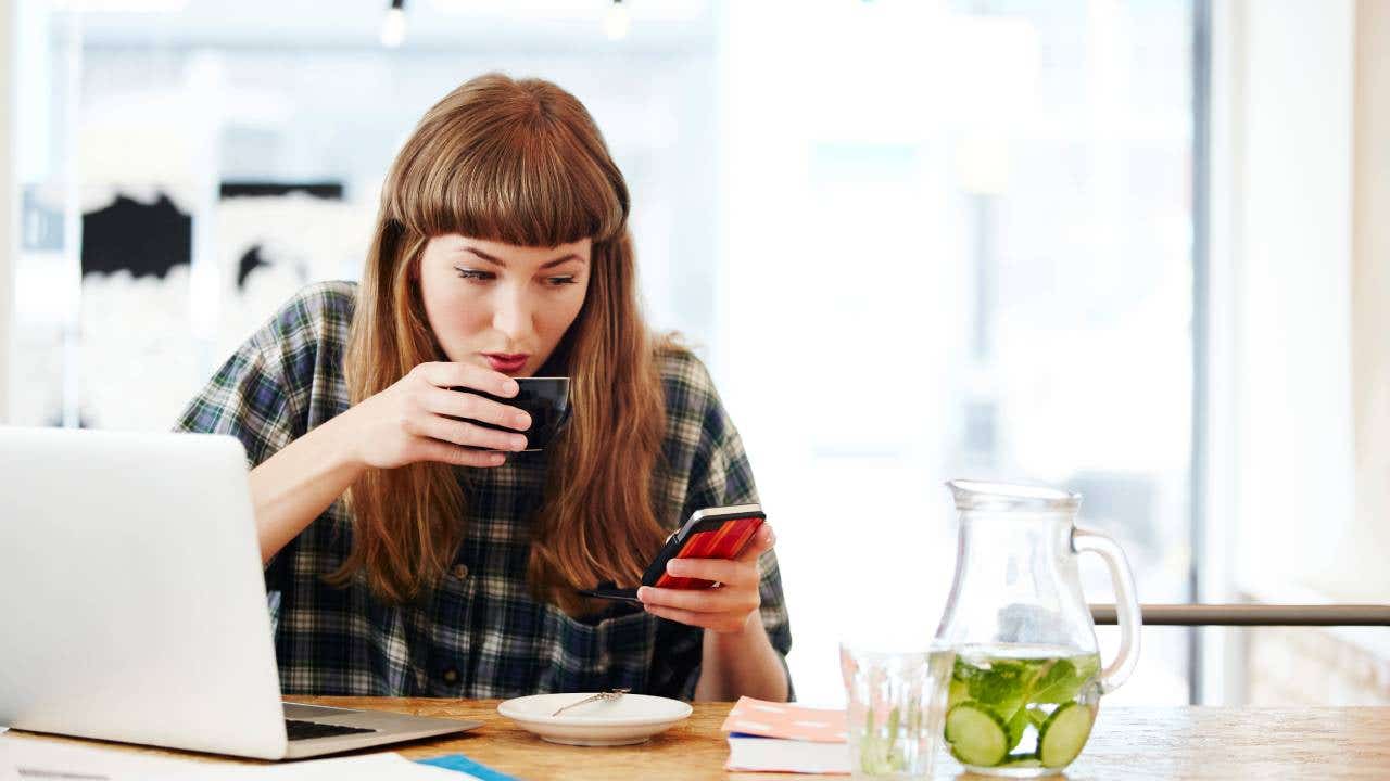 Person working at table with computer, drinking coffee and looking at smartphone