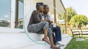 An older couple sits on the steps of a deck outside drinking coffee.