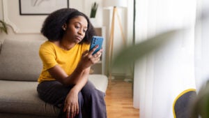 Young Black woman sitting at home looking thoughtfully at her phone.