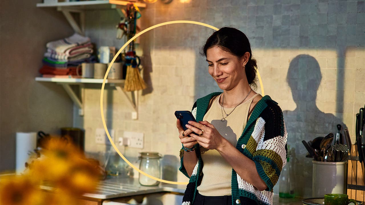 A woman stands in a sunny kitchen looking at her phone and smiling.