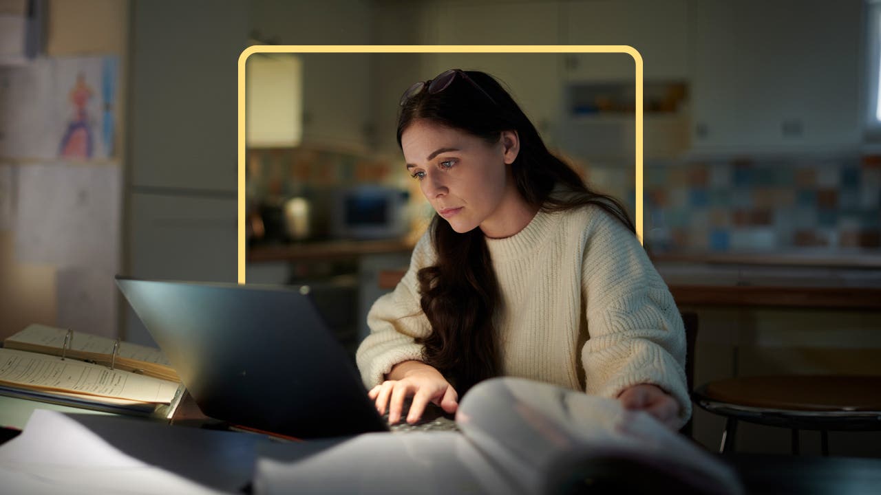 Woman with long dark hair sitting at a table working on a laptop computer that is surrounded by open binders and books.