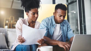 male and female sitting on a couch at home looking at paper bills and laptop screen