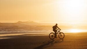 A woman goes for a bike ride at low tide on Long Beach on Vancouver Island near Tofino, British Columbia, Canada, at sunset. She wears casual clothing, a sunhat, and a backpack.