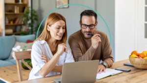 A man and a woman sit at a desk while looking at a laptop.